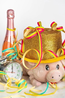 Plush pig wearing a golden hat next to an alarm clock and champagne bottle, colourful streamers,