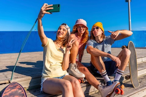 Three young, stylish friends are sitting on a wooden boardwalk, taking a selfie with a smartphone