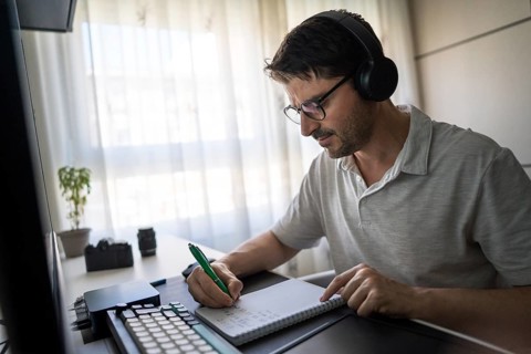 A man wearing headphones is focused on studying Mandarin online at home. He is taking notes on a