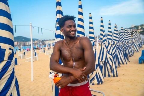 Young shirtless man standing on a sandy beach, joyfully applying sunscreen to his arm, with rows of