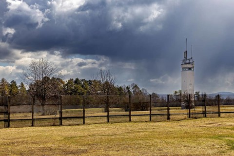 A watchtower stands behind a fence in a meadow under a cloudy sky, The Point Alpha memorial between