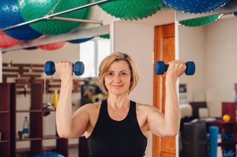 Mature woman lifting dumbbells in a gym environment, confidently looking at the camera. Her