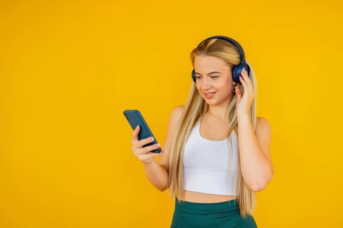 A young woman in a white top and green pants enjoys music through headphones while holding a