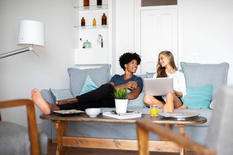 Two friends sit comfortably on a modern couch. One holds a laptop, while the other enjoys a drink,