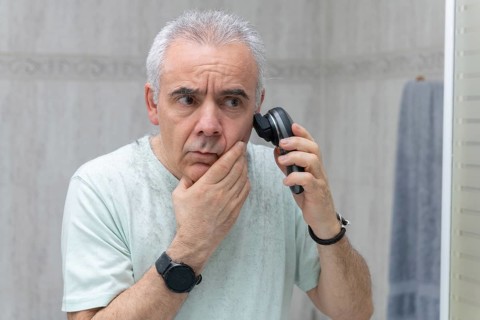 A mature man is carefully trimming his gray hair at home using an electric trimmer. He stands in