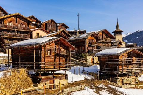 Snow-covered roofs of old farmhouses and barn storage, in the back the wooden cross and the church