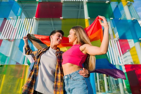 A joyful lesbian couple embraces while holding the Pride and Transgender flags. Their loving gaze