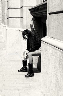 A fashionable woman wearing bold boots and patterned pants sits against a historic building.