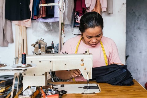 A asian seamstress focuses on sewing in a small workshop. Surrounded by tools and garments, she