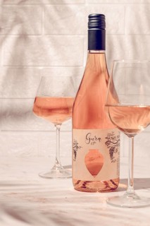 A bottle of Gura wine sits on a table beside two glasses filled with wine. The bright setting