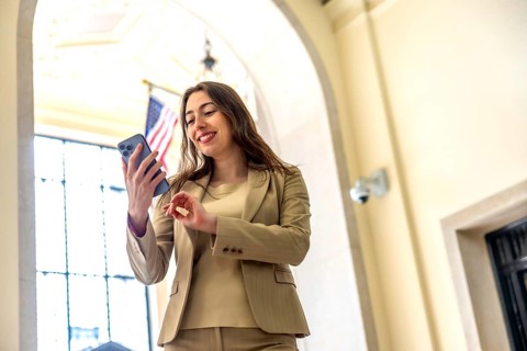 Businesswoman in a brown suit smiling as she scrolls a smartphone inside a bright, arched manhattan