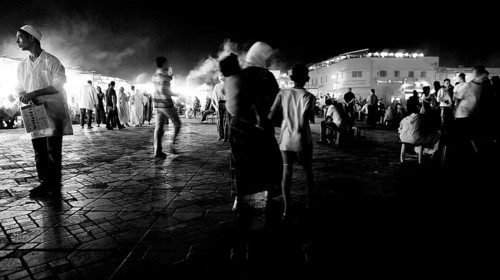Groups of people strolling on a paved black and white night market, evening food stalls at Djemaa