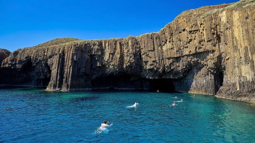 People swim in clear water in front of impressive basalt columns under blue sky, Glaronisia,
