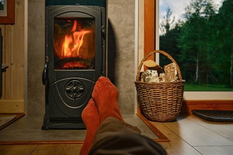 Feet in red socks are resting in front of wood stove in wooden cabin. Flames are visible, creating