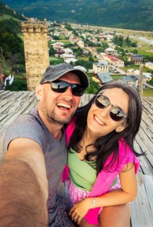 Caucasian tourist couple sit on Svan tower rooftop taking selfie watch sunset town panorama of