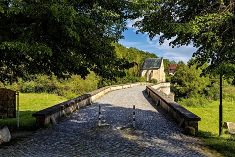 An old stone bridge leads across a quiet river to a small church in the countryside on a sunny day,