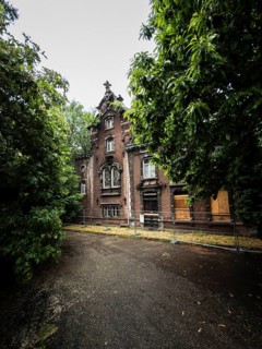 An old brick villa stands abandoned and boarded up with wooden panels behind a construction fence,