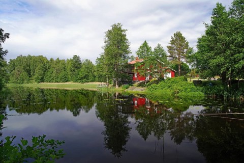 Lakeside with red house on the edge of the forest. Trees are reflected in the water. Peaceful,