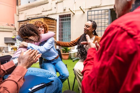 Diverse individuals sitting in a circle during group therapy, with two women hugging in an