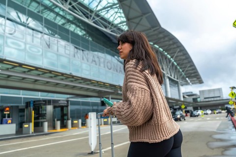 Young woman standing near the curb with luggage, holding a phone, and looking to the side, waiting