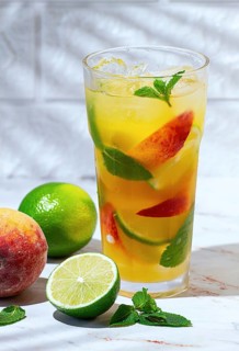 A tall glass filled with a drink made from lime and peach sits on a marble surface. Fresh mint