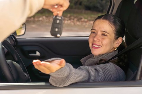 Smiling young woman looking at car key being handed to her through window, car interior,