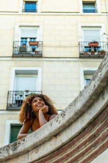 Young happy woman with curly hair smiling and looking up, leaning over a wall in an urban setting,