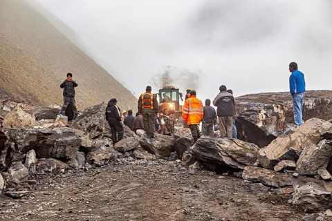 People work together on a mountainous, foggy road with a tractor in the background, rockslide on