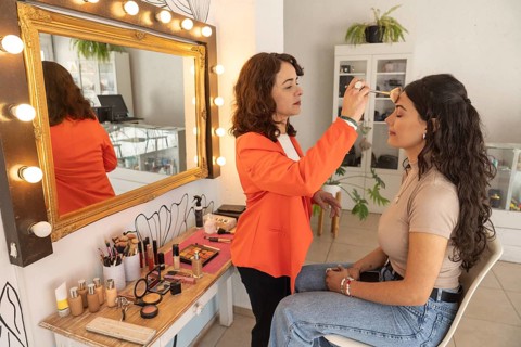 Focused makeup artist applies eyeshadow to a young client while both are reflected in an