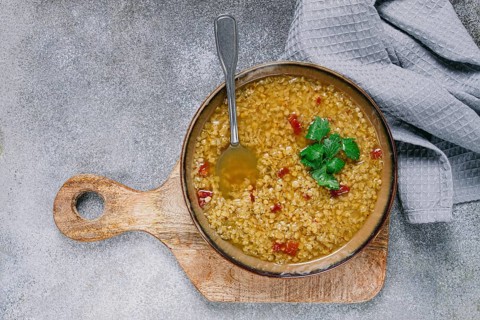 Savory lentil soup garnished with fresh herbs sits in a wooden bowl on a textured surface,