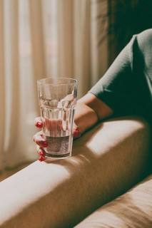 A person holds a clear glass filled with water while sitting on a couch. Sunlight streams through a