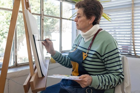Older female artist applies brushstrokes to a canvas while holding a watercolor palette and working