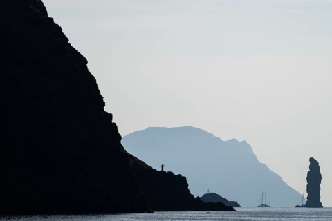 Dramatic cliffs and silhouette of a towering rock pin, behind it the island of Alicudi, a sailboat