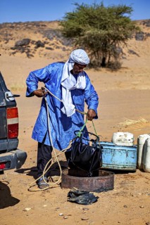A man in a blue robe deals with a water container in a desert-like environment, Tuareg draw water