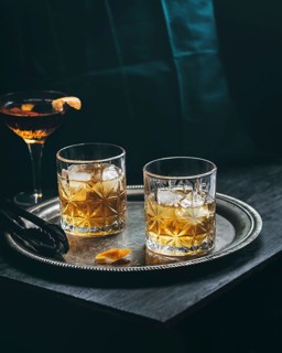 Two whiskey glasses filled with ice rest on a silver tray alongside a cocktail featuring a vibrant