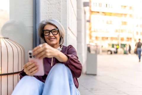 Senior woman with grey hair and glasses sitting casually on a sidewalk, smiling while using her