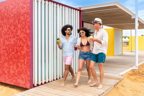 Diverse friends walking barefoot on a sunny beach boardwalk, laughing and sipping cold drinks