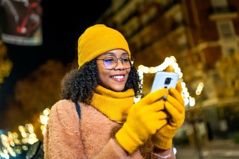 Smiling young woman wearing yellow winter clothes and gloves using smartphone in a city street at