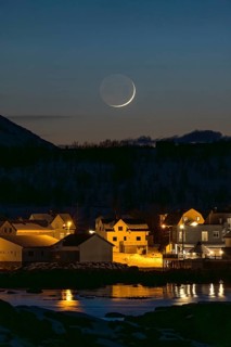 A serene nordic village is bathed in the soft ashen light under a crescent moon. The calm water