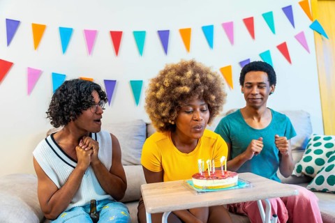 African young woman with curly hair blowing candles on birthday cake sitting on the sofa with