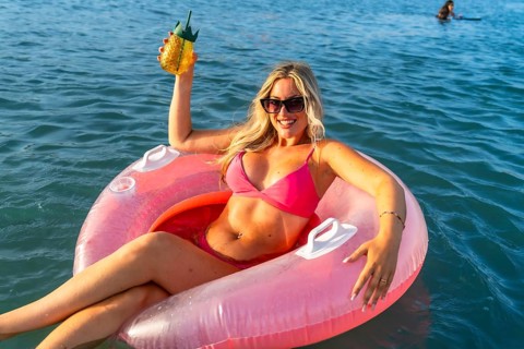 Young woman smiling while floating in the ocean on a pink inflatable ring, holding a tropical drink