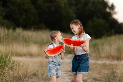 Two children smiling and holding watermelon slices outdoors on a summer day. They stand in a grassy