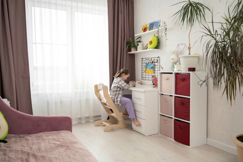 A young girl sits barefoot at her desk in a bright, organized bedroom, drawing on a digital tablet.