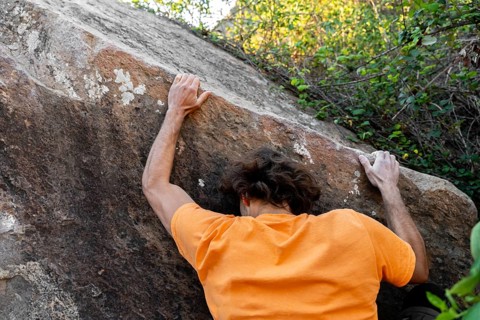 A climber in a vibrant orange shirt scales a rugged boulder in a lush natural setting, showcasing