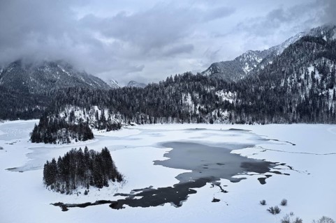 Ice-covered and snow-covered lake in wintry mountain landscape, cloudy, snow-covered trees,