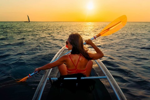 Silhouetted pretty sporty fit woman paddles clear kayak during vibrant Zanzibar sunset. Back view