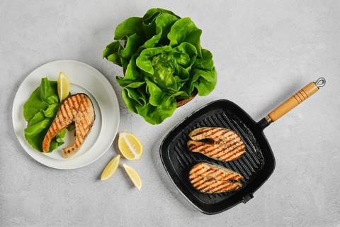 Grilled salmon fillets rest on plate with salad leaves. Griddle pan with grilled steaks is nearby.