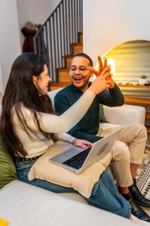 Diverse couple high fiving on a cozy living room couch, celebrating a successful remote work win