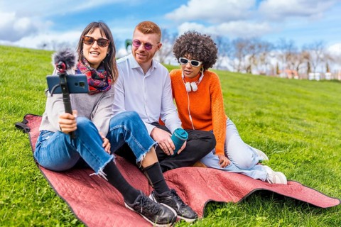 Three content creators filming a video blog in a park using a smartphone with external microphone,