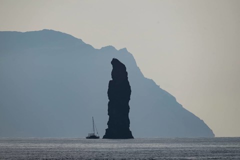 Silhouette of a towering rock needle, behind it the island of Alicudi, a sailboat in the calm sea